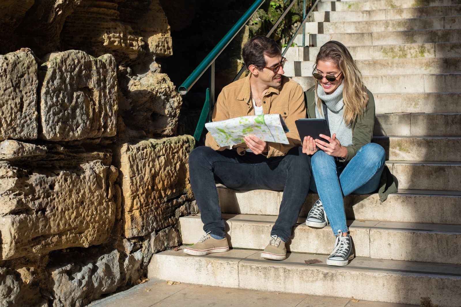 A smiling couple sitting on old stone stairs with a map and tablet, asking questions about a personal cruise planner